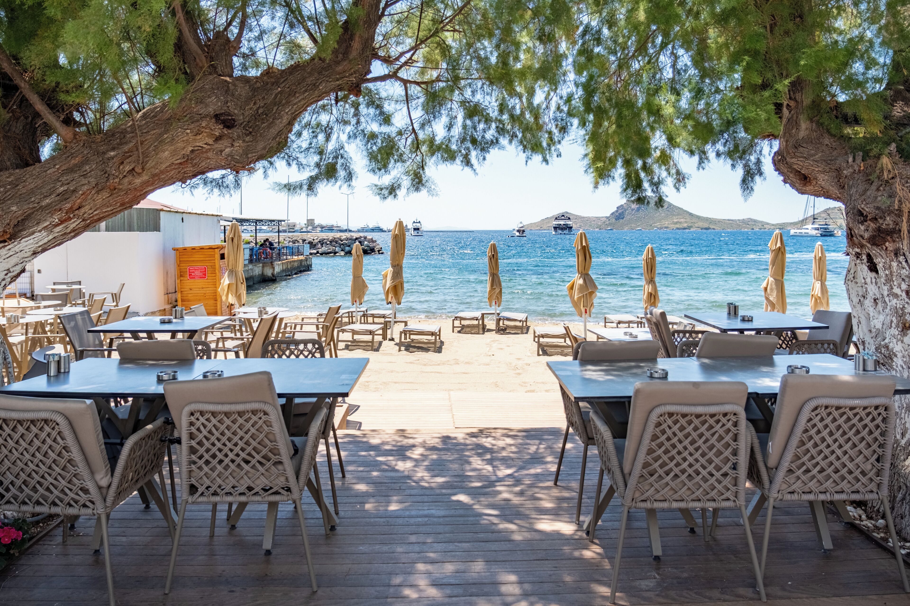 A seaside restaurant with outdoor seating, a view of the sea, and a dock in the background.