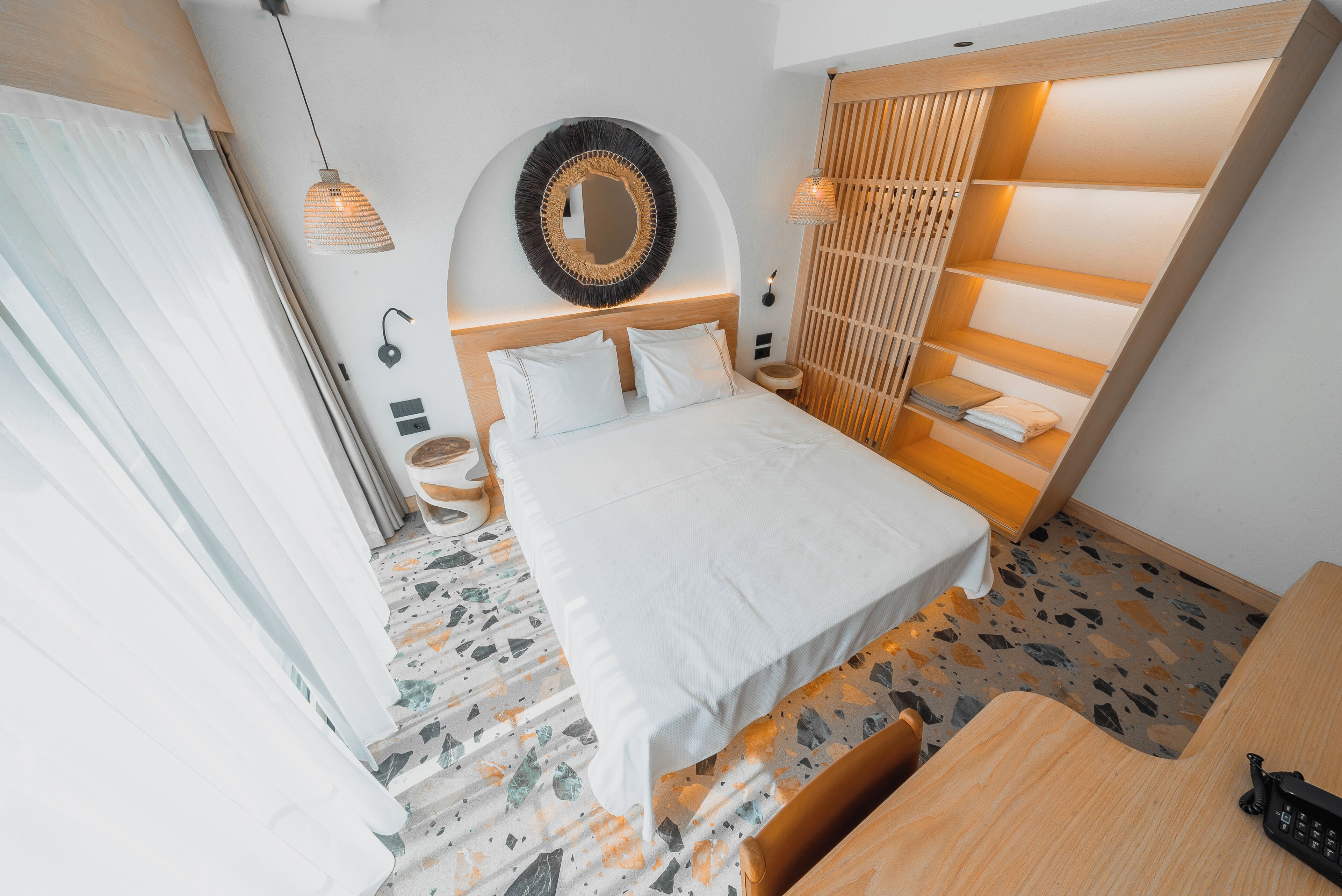 A modern hotel room with a bed, a round mirror, a wooden headboard, a desk, and a small seating area.