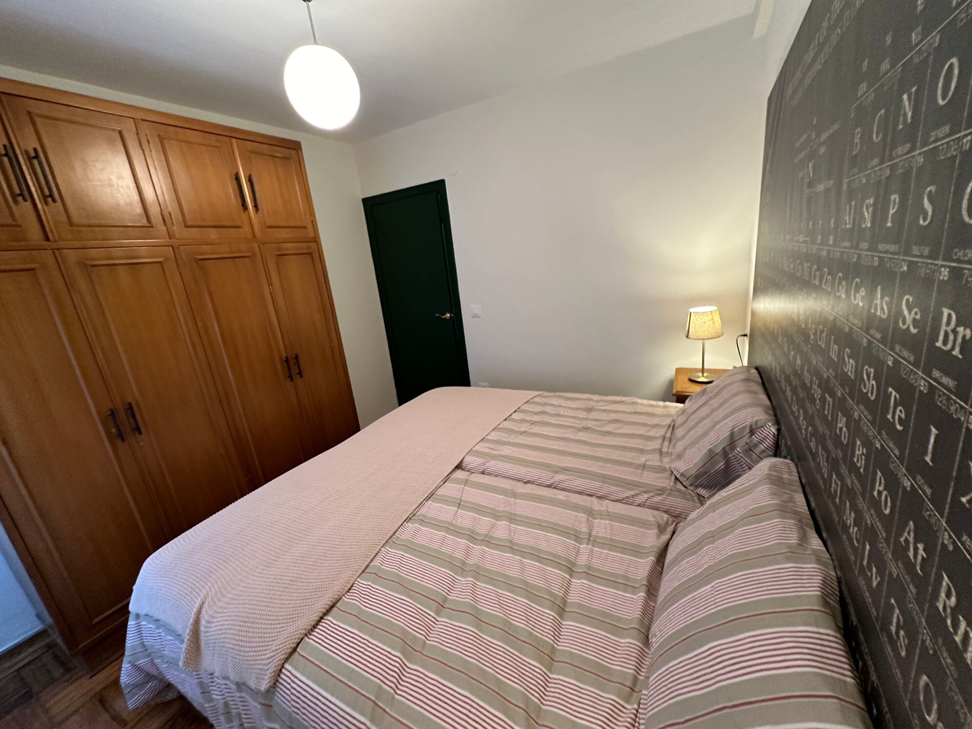 Une chambre à coucher avec un lit, une armoire en bois, une porte verte, un mur orné d’un tableau périodique et une lampe de chevet.