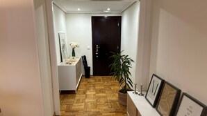 Un pasillo con suelo de madera, una puerta oscura al final, una consola blanca con un florero y una vela, y texto enmarcados en la pared.