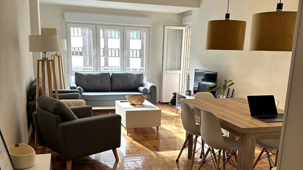 Una sala de estar moderna con un sofá, una mesa de comedor con sillas y un ventanal con vistas a edificios.