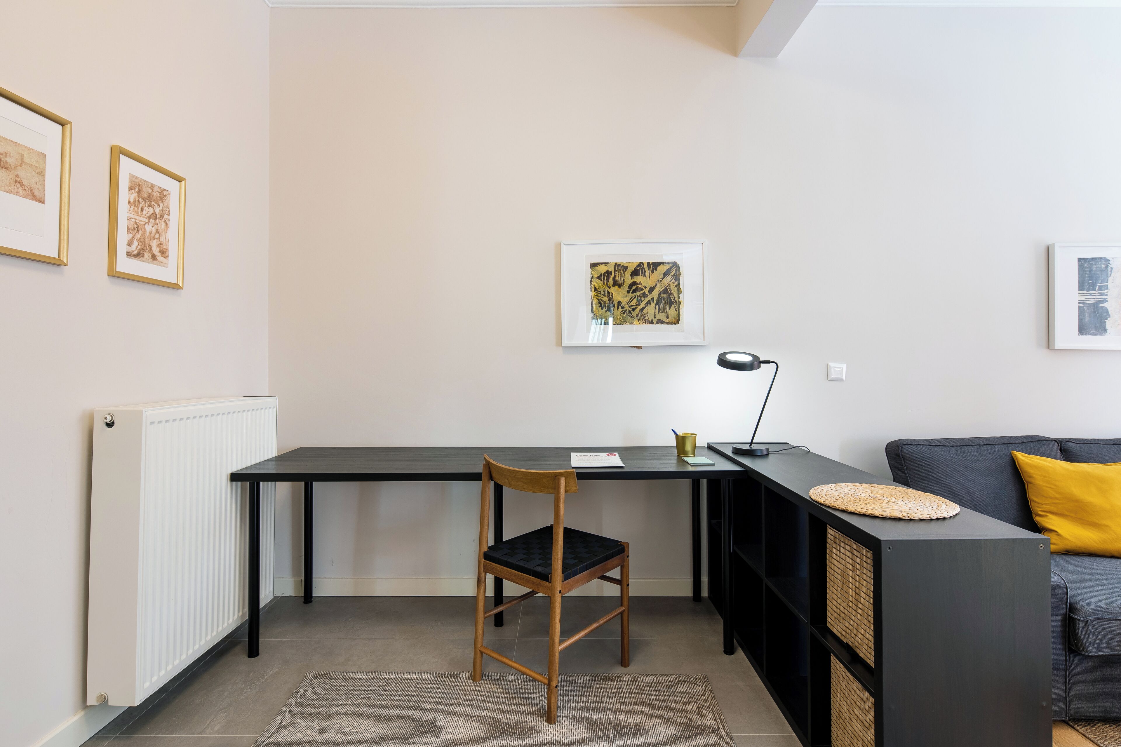 A modern living room with a black desk, a wooden chair, a grey sofa with a yellow pillow, and framed artwork on the walls.