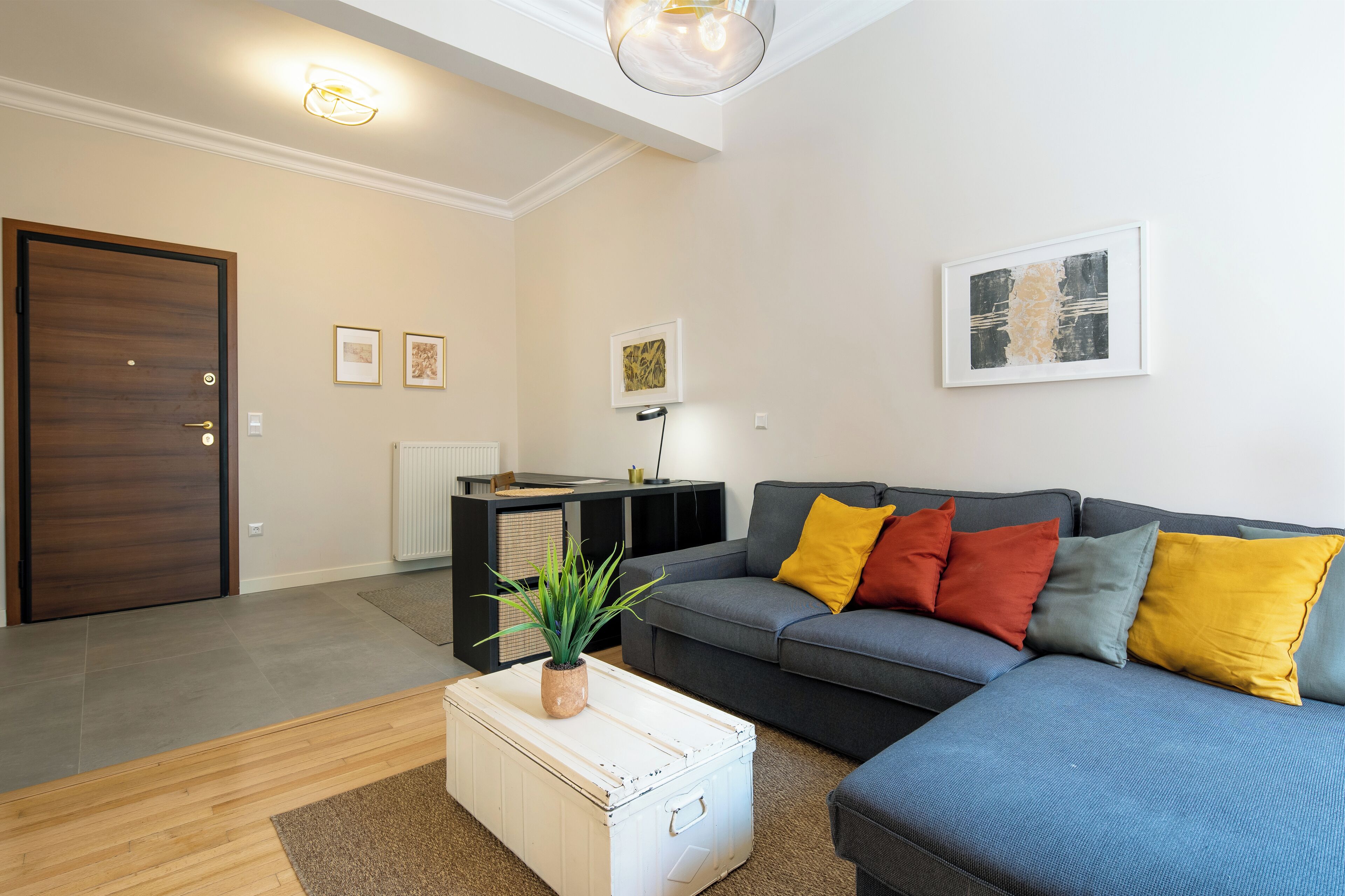 Un salon moderne avec un canapé gris, des coussins colorés, une table basse blanche agrémentée d’une plante et des œuvres d’art accrochées aux murs.