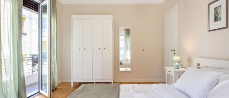 A hotel room with a bed, a wardrobe, a bedside table with a lamp, a mirror, and a window with curtains.
