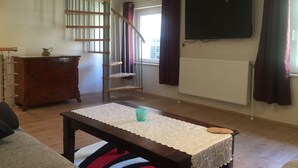 Una sala de estar con una escalera, una mesa de madera con mantel de encaje, un televisor montado en la pared y un radiador.