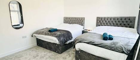 Ein Schlafzimmer mit zwei Betten, jeweils mit grauem Kopfteil und grauer Bettdecke. An der Wand hängt ein Spiegel und ein kleiner Tisch mit einer Lampe steht daneben.