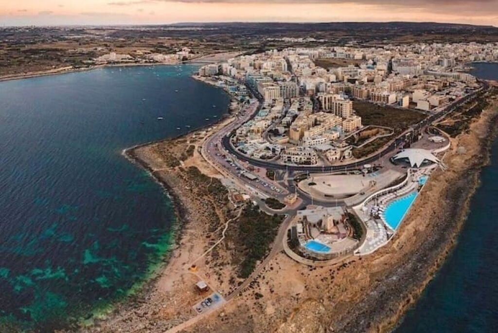 An aerial view of a coastal city with a marina, a large central building complex, and a circular park with a pool.