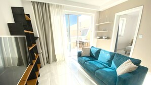Een moderne woonkamer met een blauwe bank, een flatscreen televisie en een eethoek zichtbaar door een glazen deur.