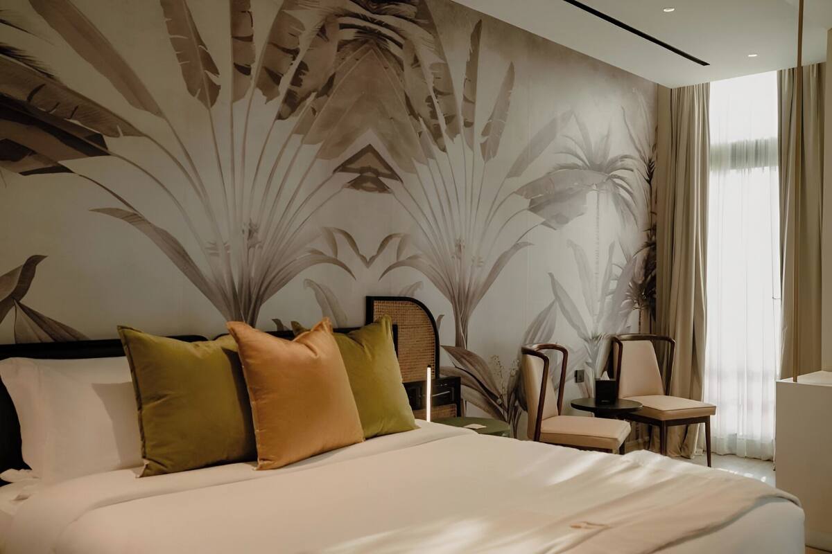 A modern hotel room with a large bed, a chair, a small table, and a large window with curtains.