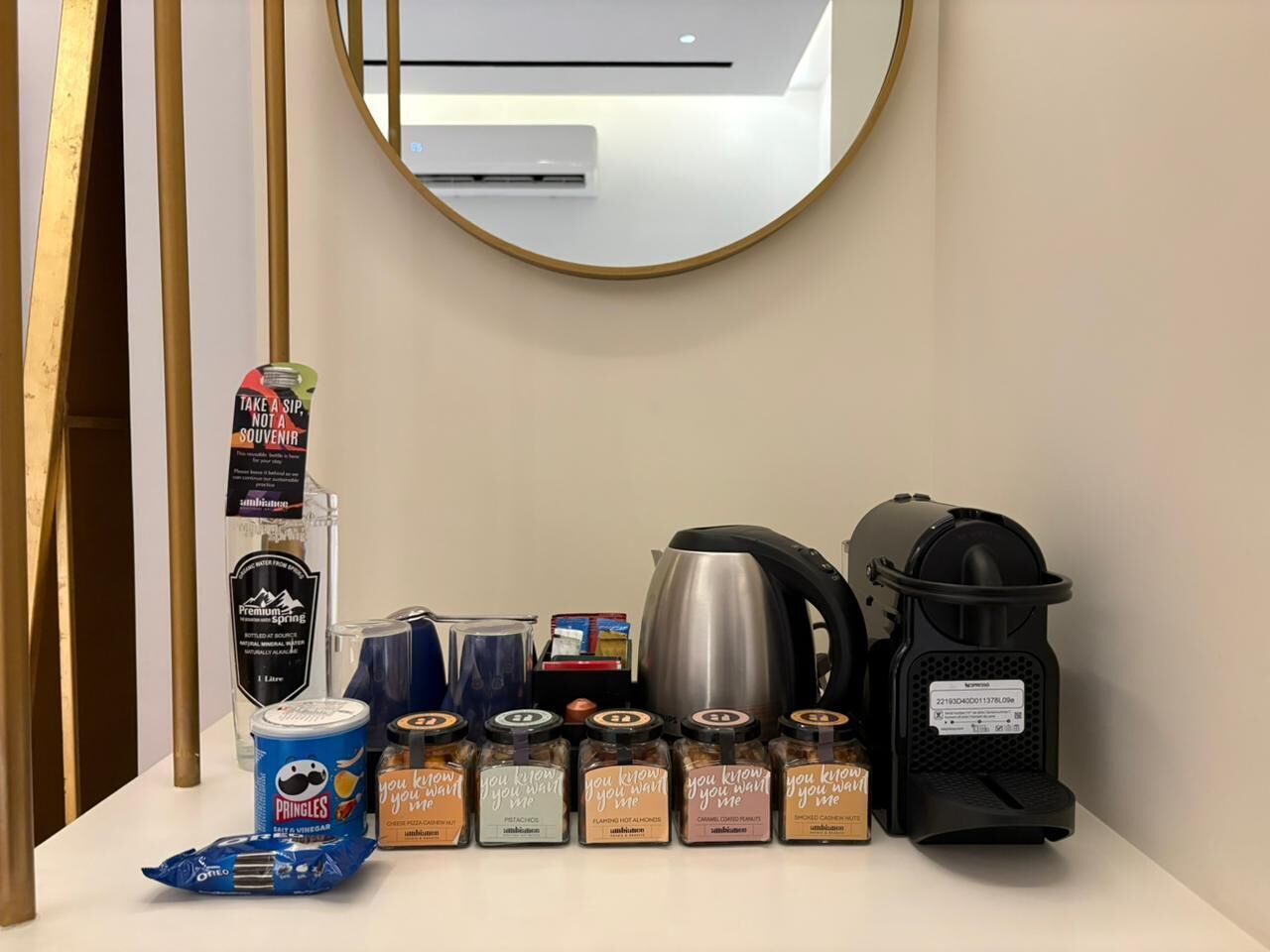 A hotel minibar setup with a bottle of water, a can of Pringles, a coffee maker, a kettle, and several jars of condiments.