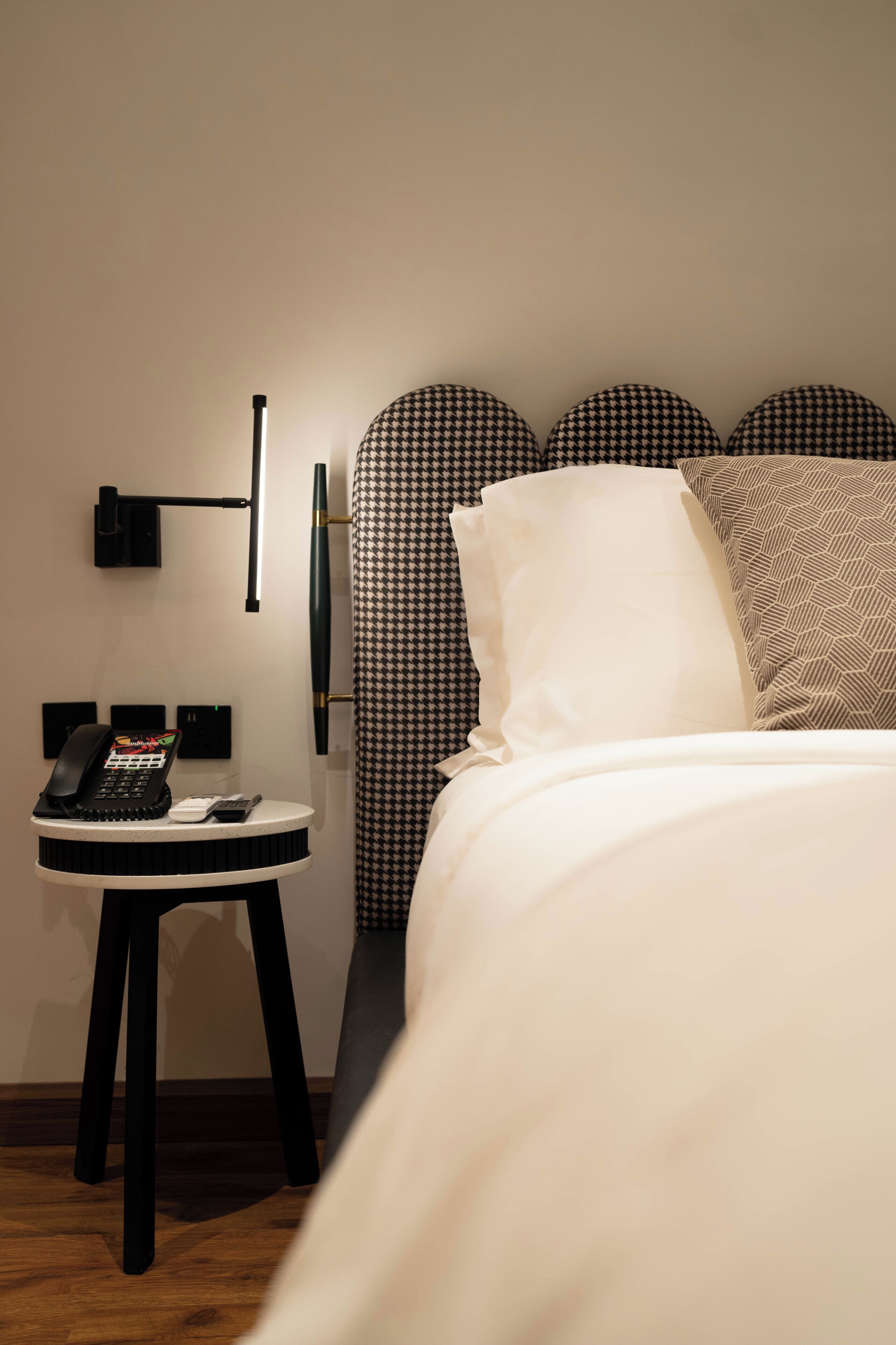 A hotel room with a bed, a bedside table with a phone, and a wall-mounted lamp.
