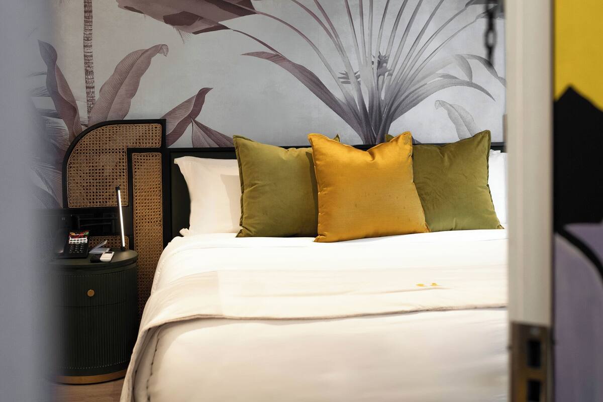 A bedroom with a bed, pillows, a nightstand, and a wall with a large plant mural.