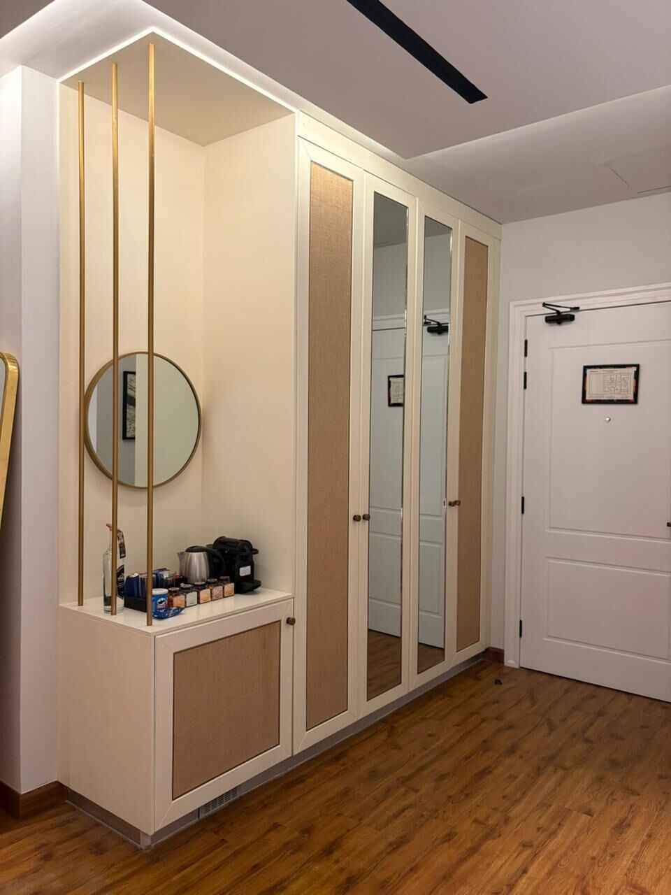 Una habitación con piso de madera, una puerta blanca, un armario blanco con espejo y una pequeña mesa con tetera y tazas.