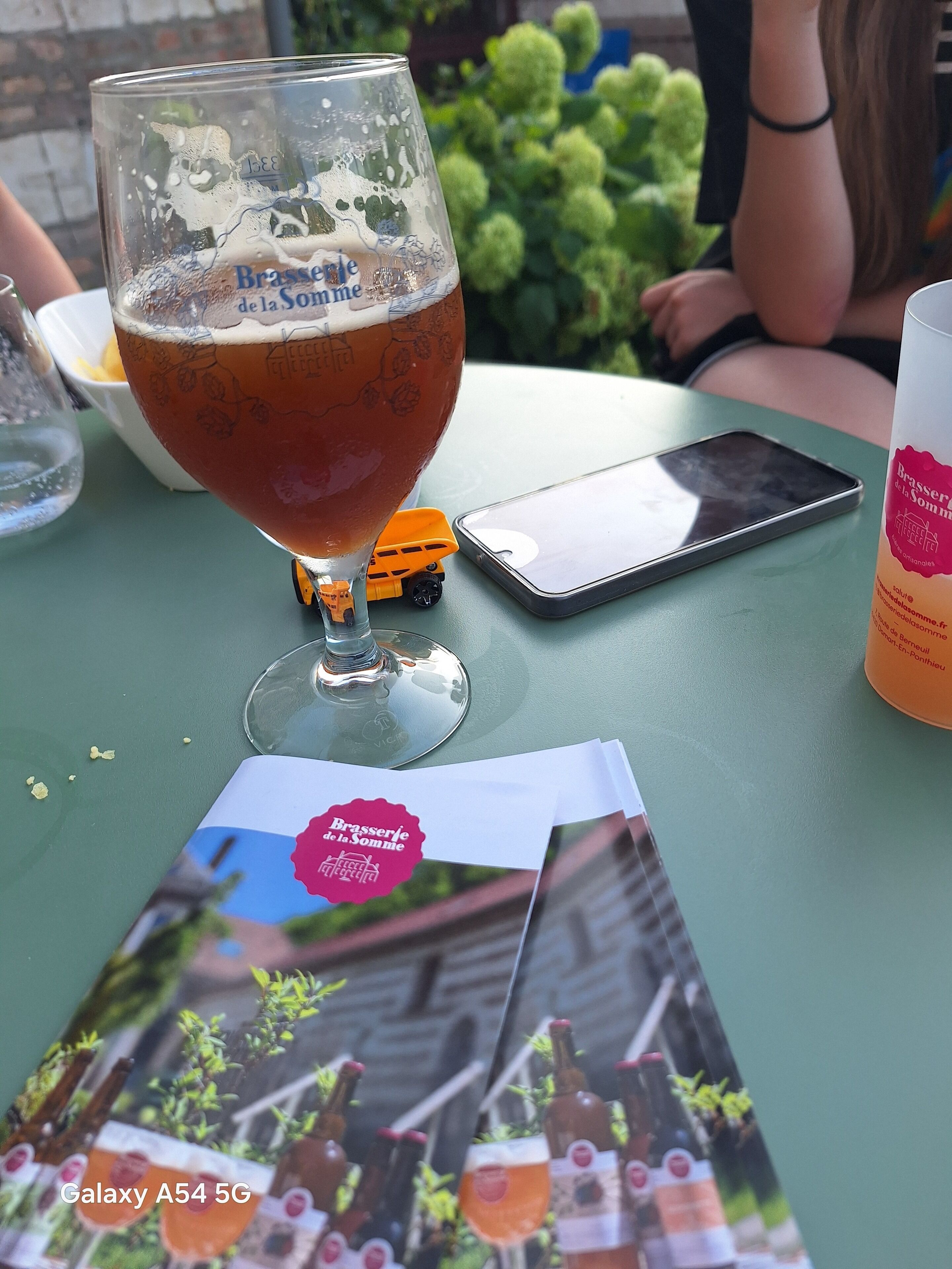 Un verre de bière sur une table, accompagné d’une brochure, d’un téléphone intelligent et d’une petite voiture jouet.
