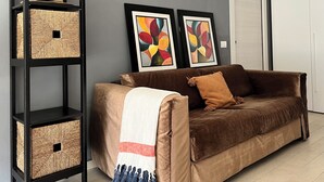 A brown sofa with a white and red blanket, a blue tassel throw, and a brown cushion. A black shelving unit with woven baskets is to the left. Two abstract paintings hang on the wall above the sofa.