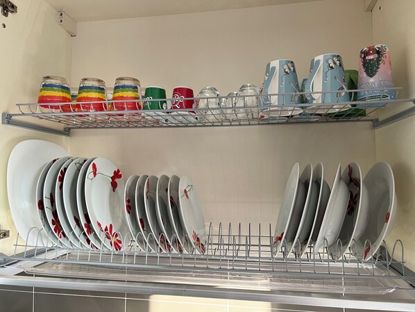 Una cocina con un escurreplatos lleno de platos y tazones, y estantes superiores con diversos recipientes y utensilios de cocina.