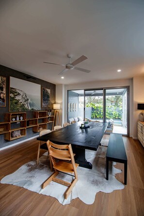 Zona pranzo moderna con un tavolo grande, sedie in legno e una panca. Si vedono scaffali con oggetti decorativi e una ventola a soffitto.