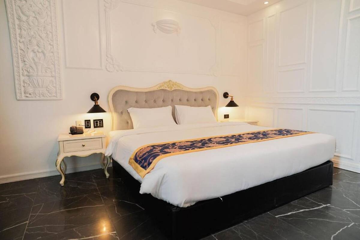 Un lit bien fait, recouvert d’une couvre-lit blanche et dorée, est flanqué de deux tables de chevet sur lesquelles reposent des lampes. La pièce est dotée d’un sol en marbre foncé et de murs blancs.