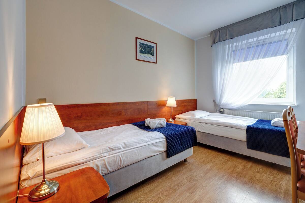 A hotel room with two single beds, a wooden headboard, a nightstand with a lamp, a framed picture on the wall, and a window with sheer curtains.