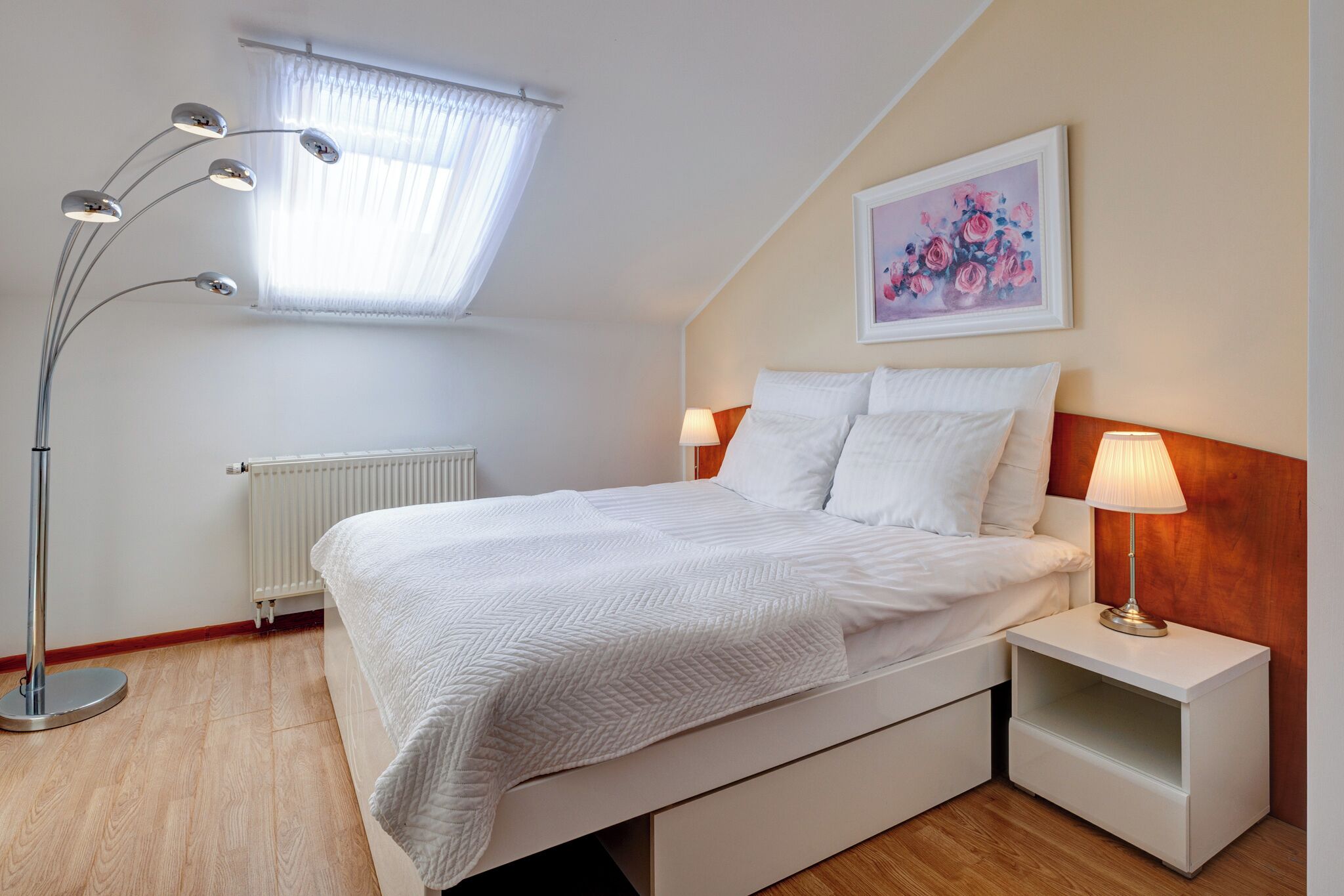 A bedroom with a bed, bedside lamps, a radiator, a floor lamp, and a painting on the wall.