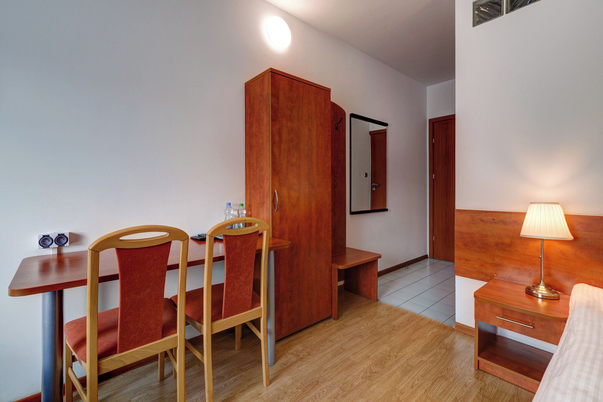 A compact hotel room with a wooden desk, two chairs, a wardrobe, a bedside table with a lamp, and a bed.