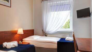 A hotel room with a bed, a bedside table with a lamp, a TV mounted on the wall, and a window with sheer curtains.