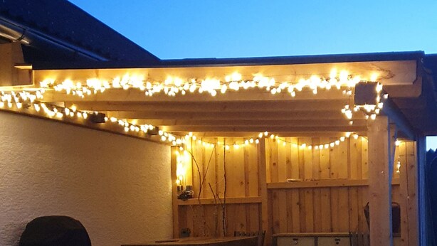 A covered outdoor patio with wooden furniture, string lights, and a barbecue grill.