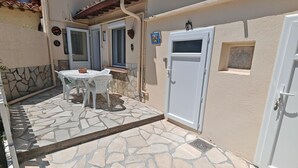 Une terrasse dallée de pierres, avec une table et des chaises blanches, une porte blanche et une petite fenêtre.