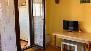 Una habitación con suelo de madera, un televisor sobre una mesa de madera, un banco blanco y una puerta que da a otra habitación con cortinas estampadas.