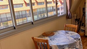Un comedor con una mesa cubierta por un mantel azul y blanco estampado, dos sillas de madera y un ventanal con vistas a edificios residenciales.