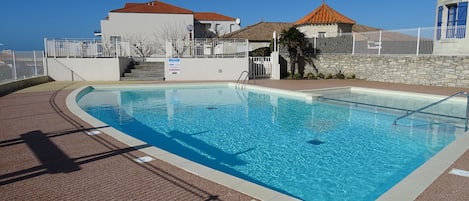 En rektangulær swimmingpool med klart, blåt vand, omgivet af en flisebelagt terrasse. Der er trapper, som fører op til en bygning med røde tagsten og en hvid facade. Et hvidt hegn omkranser poolområdet.