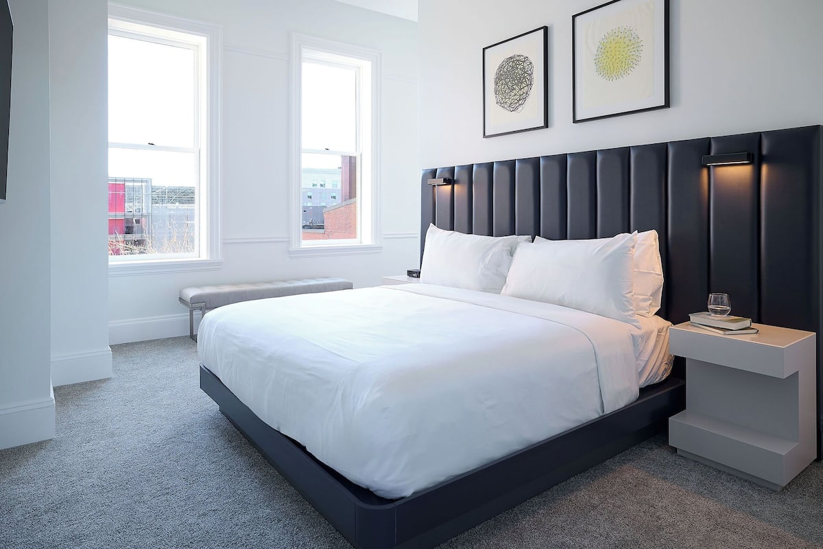 Ein modernes Schlafzimmer mit einem großen Bett, zwei Nachttischen und Blick auf die Stadt durch ein Fenster.