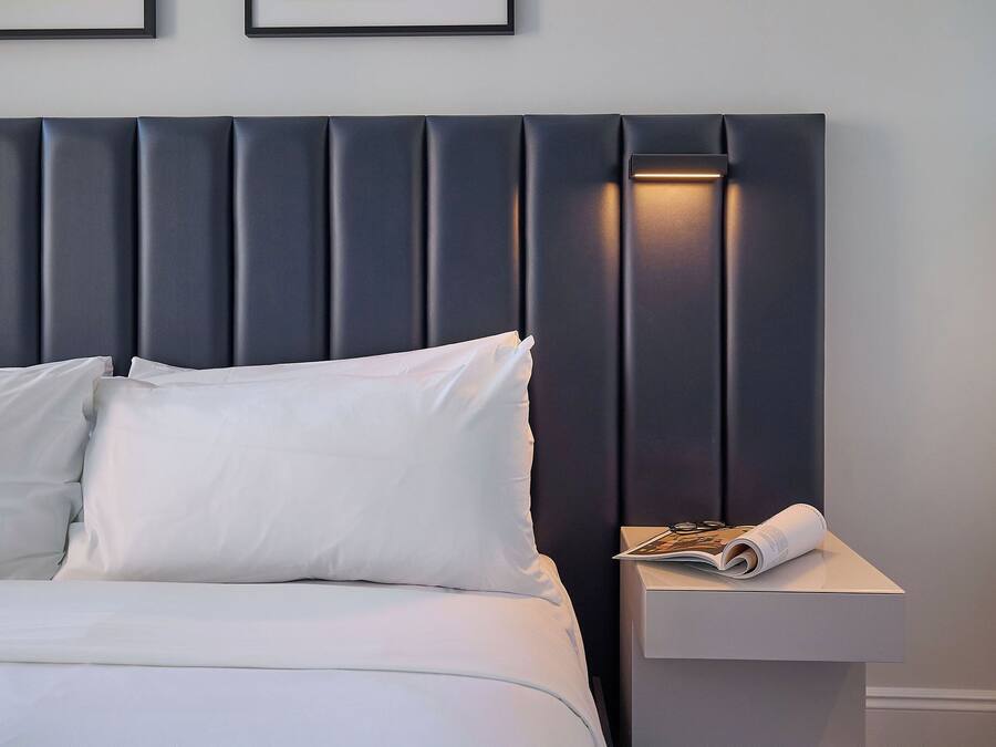 Una habitación de hotel moderna con una cama grande, ropa de cama blanca, un cabecero oscuro y una mesita de noche con una revista.