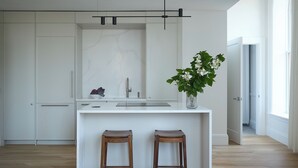 Una cucina moderna con mobili bianchi, una parete rivestita in marmo, due sgabelli in legno e una pianta in vaso.