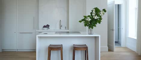 Uma cozinha moderna com armários brancos, painel de backsplash em mármore, dois banquinhos de madeira e um vaso com planta.