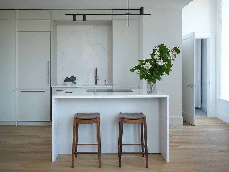 A modern kitchen with white cabinetry, a marble backsplash, two wooden stools, and a potted plant.