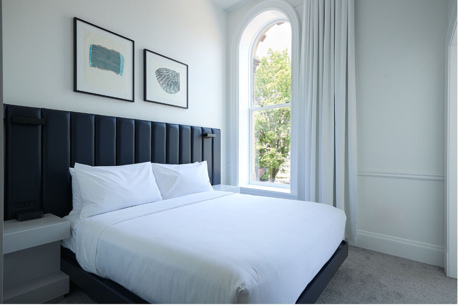 A neatly made bed with white linens in a room with a dark headboard, a window with curtains, and two framed artworks on the wall.