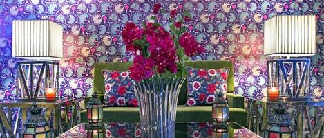 A room with a patterned wallpaper, a green sofa with floral cushions, a glass table, and two wall-mounted lanterns.