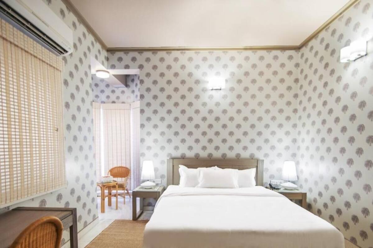 Une chambre d’hôtel avec un grand lit, deux tables de chevet avec des lampes, un petit bureau avec une chaise et une fenêtre avec des stores.