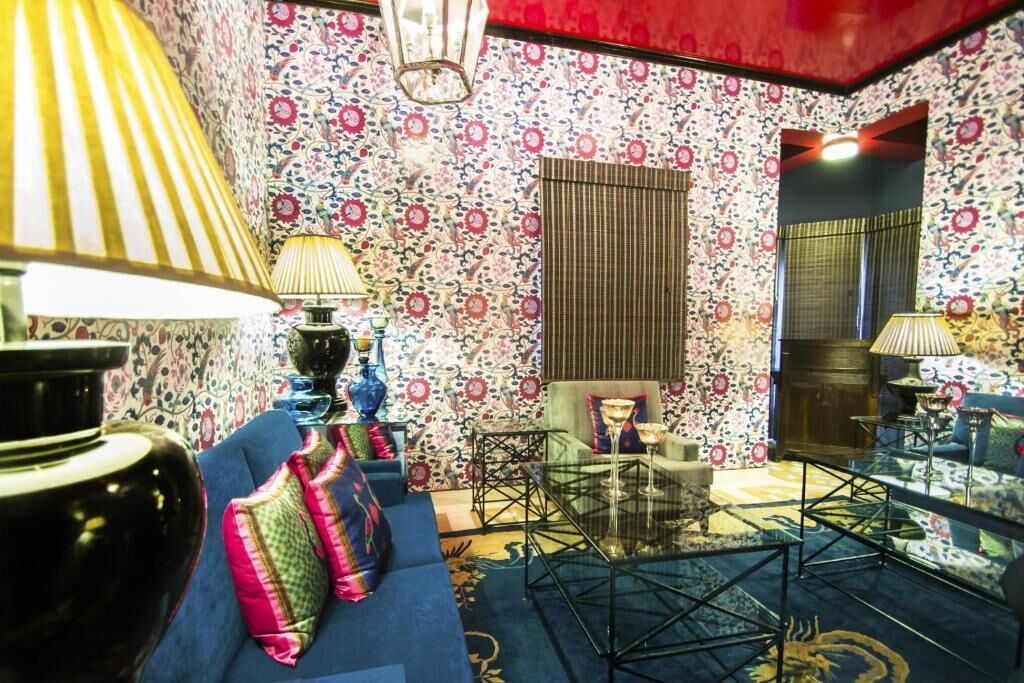 Una habitación con papel tapiz floral, un sofá azul, una mesa de centro de vidrio y una lámpara.