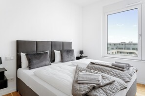 A modern bedroom with a large bed, grey bedding, and a view of buildings through a window.