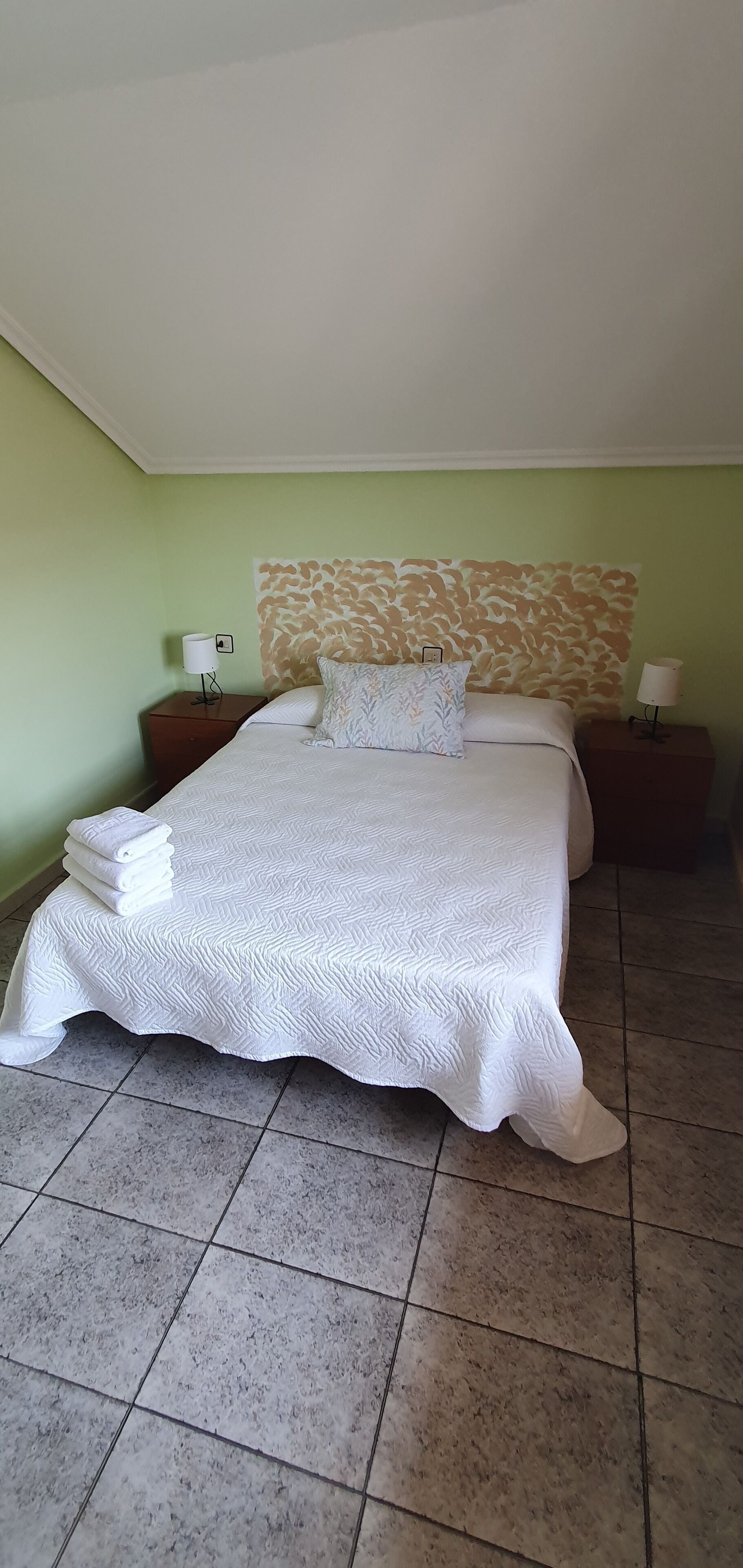 Una cama bien hecha con una colcha estampada, dos almohadas y un cabecero con diseño de hojas. Hay dos mesitas de noche con lámparas, y una pila de toallas dobladas sobre la cama.