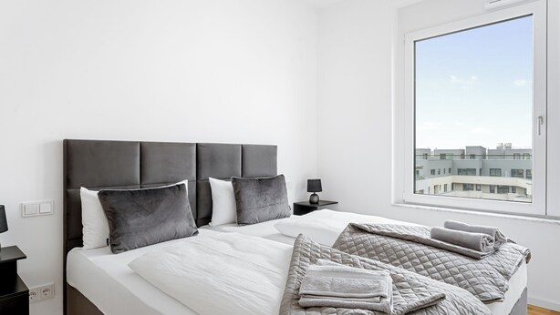 A modern bedroom with a large bed, grey bedding, and a view of buildings through a window.