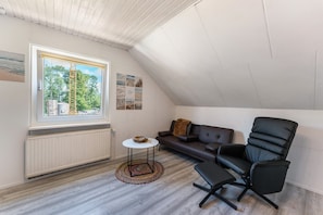 A room with a sloped ceiling, a window with a view of trees, a black leather sofa, a black office chair, and a small round table with a bowl. - Ostseeclick Damp – Beachside Apartment with 2 Free E-Bikes (Damp)