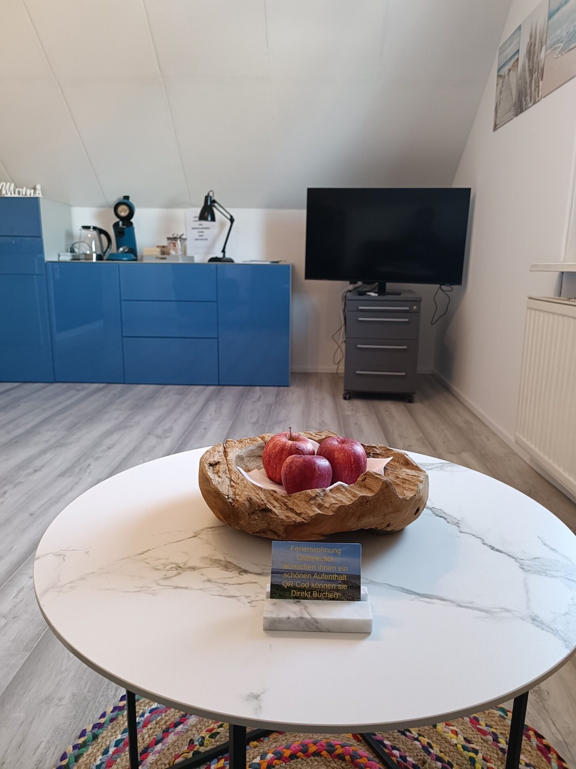 A round table with a marble top, a wooden bowl with apples, and a sign in German.