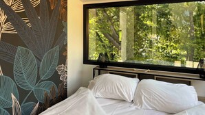 Un dormitorio con un ventanal grande, una cama con ropa de cama blanca y una pared con diseño de hojas.