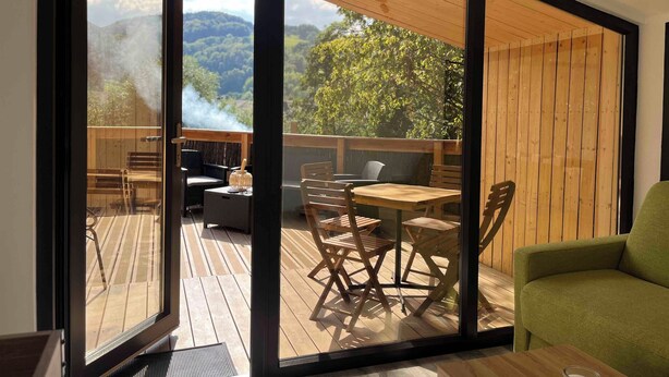 Una habitación moderna con un amplio ventanal corredera que da a una terraza exterior con mobiliario de madera y vistas a un paisaje montañoso.