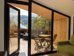 A modern room with a large sliding glass door leading to an outdoor deck with wooden furniture and a view of a mountainous landscape.