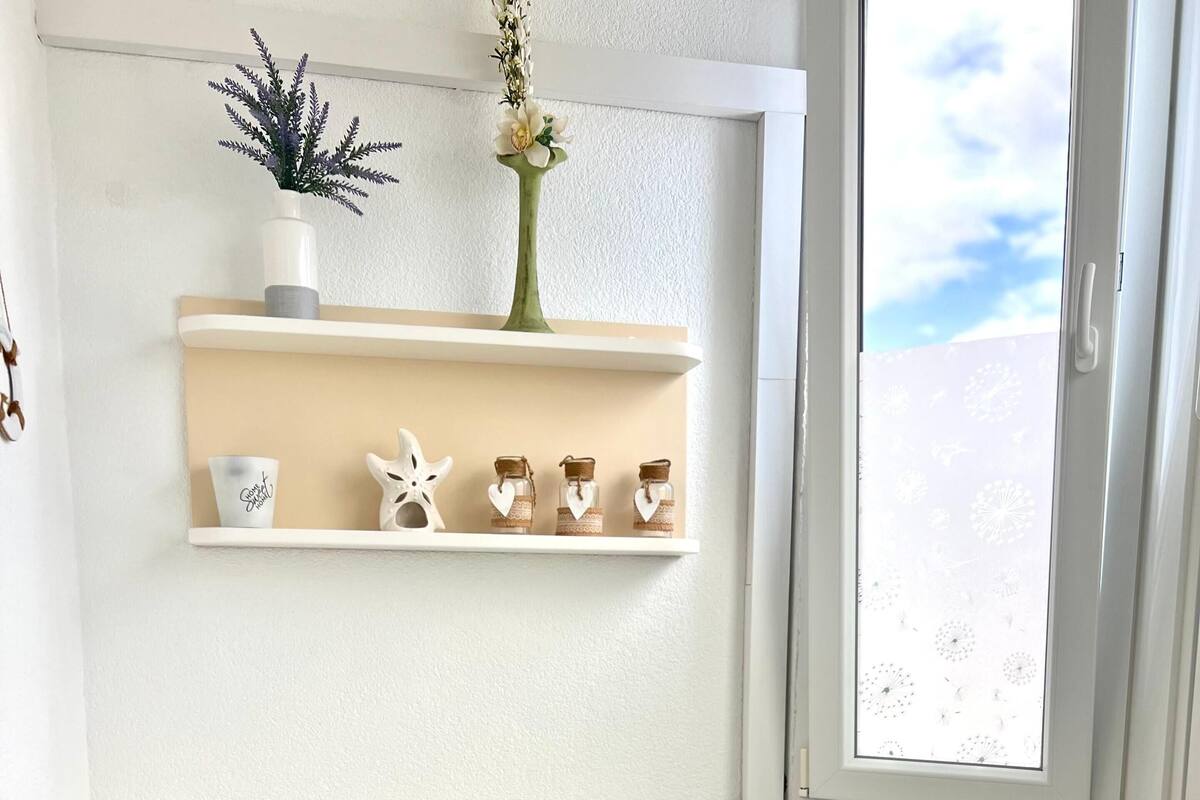 A dining area with a white table and chairs, a shelf with decorative items, and a window with a view of the sky.