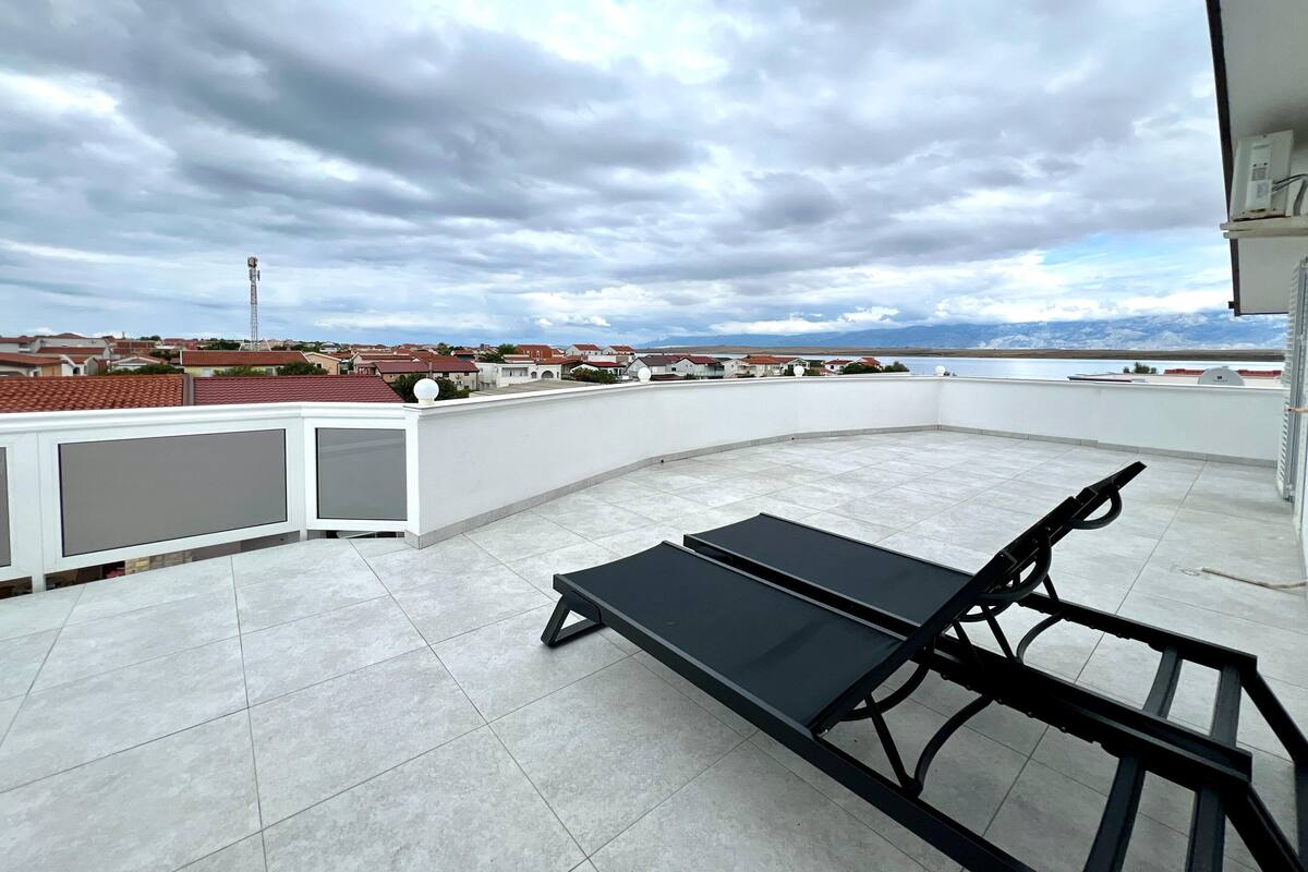 A rooftop terrace with a black lounge chair, a white wall, and a view of a residential area and mountains in the distance.